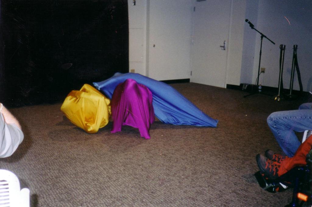 Three figures in bright pink, yellow and blue sacks ooze over each other.