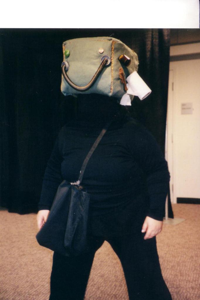 A figure in black wearing a crossbody bag with a cube for a head featuring faces made from random objects such as a spring, batteries and a toilet paper tube.