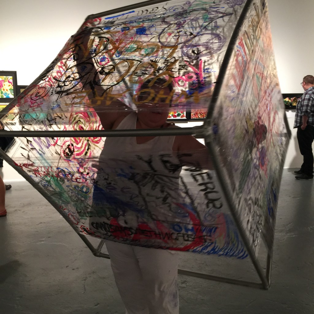 Maia wearing white inside a graffiti covered PVC cube, arms up holding the cube on an angle.