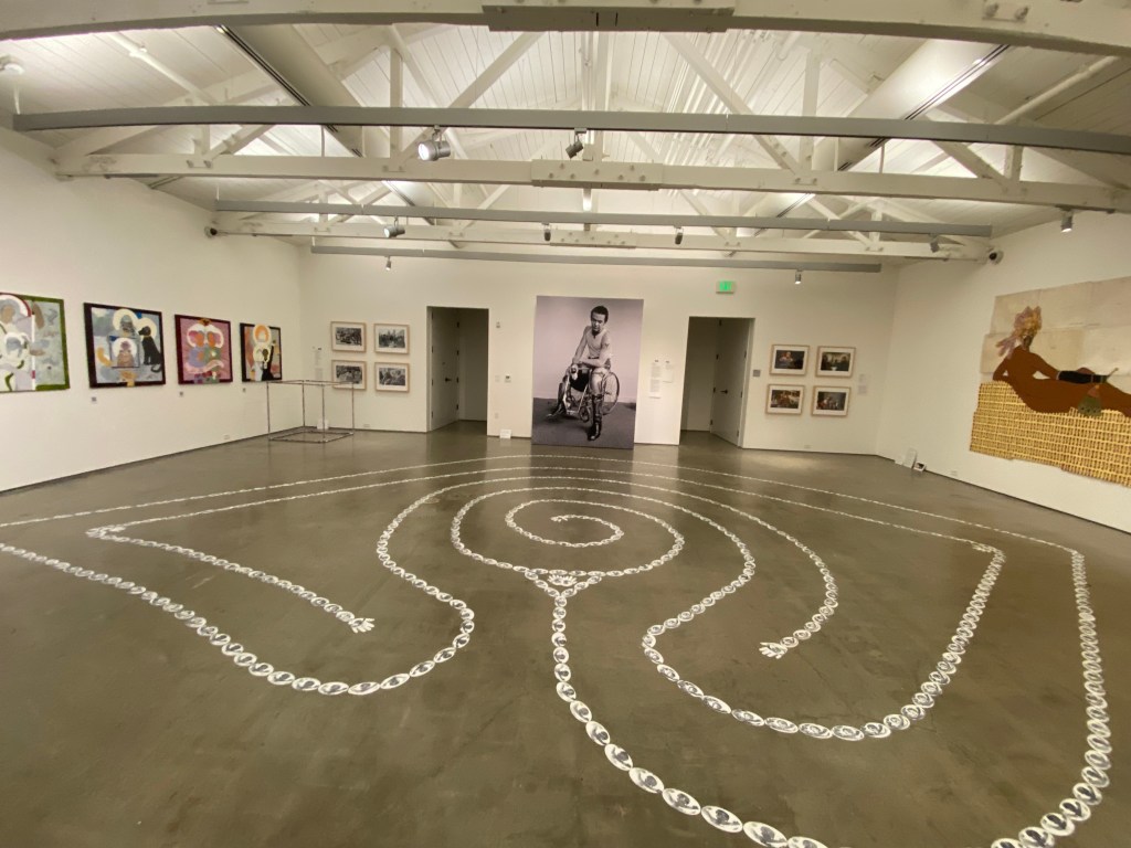 Eye shaped labyrinth made of stamped images of Maia's eye in a gallery space featuring a large-scale photo of a man ina wheelchair holding a beer.