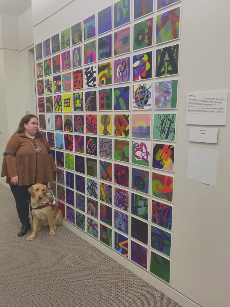 Maia and her guide dog standing next to a wall of 100 colorful squares in a grid pattern.