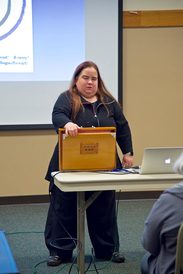 Maia standing behind a table playing a shruti box.  There's a laptop to her left and the corner of a projection to her right.